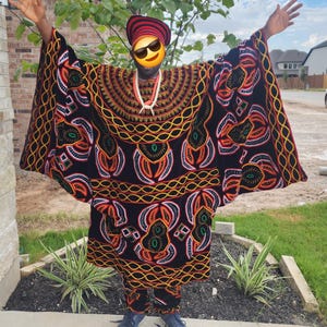 May include: A traditional African outfit in black with intricate patterns in orange, yellow, and green. The ensemble includes a long, flowing robe and matching pants. The person is also wearing a red and black striped hat and blue sneakers.