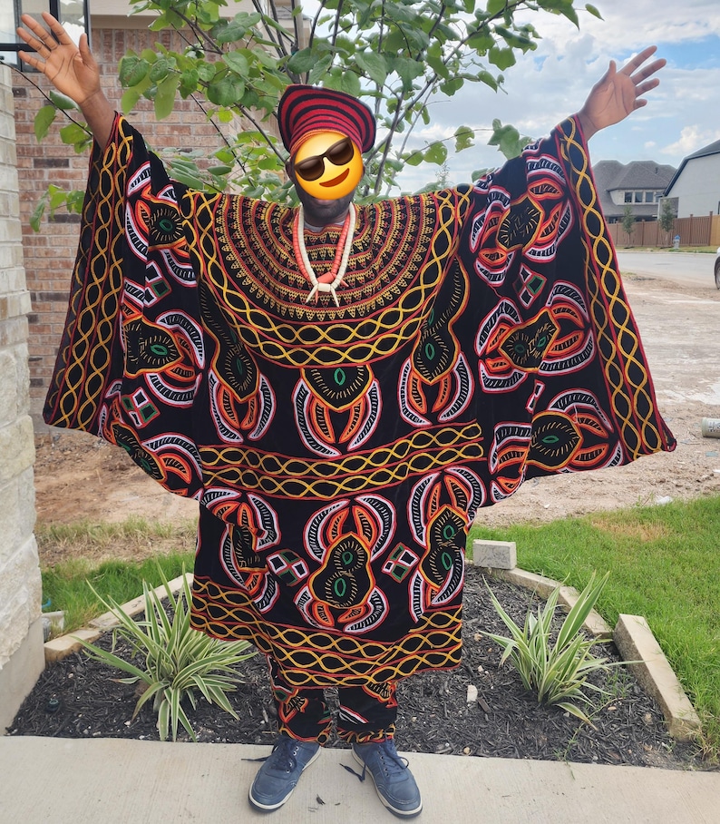May include: A traditional African outfit in black with intricate patterns in orange, yellow, and green. The ensemble includes a long, flowing robe and matching pants. The person is also wearing a red and black striped hat and blue sneakers.
