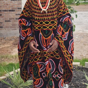May include: A person wearing a traditional African outfit. The outfit includes a black robe with colorful geometric patterns in orange, yellow, and white. A matching hat and necklace complete the ensemble. The person is also wearing blue sneakers.