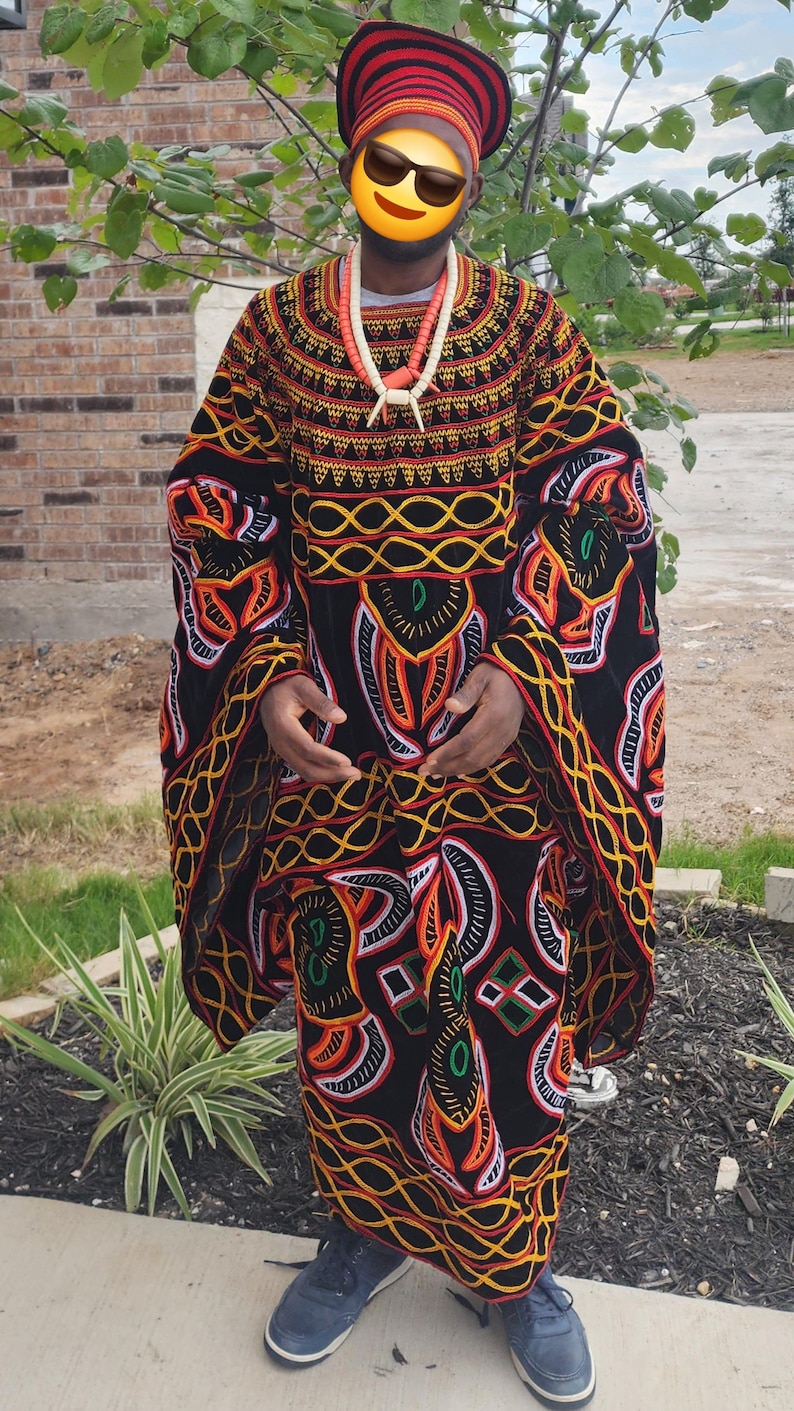 May include: A person wearing a traditional African outfit. The outfit includes a black robe with colorful geometric patterns in orange, yellow, and white. A matching hat and necklace complete the ensemble. The person is also wearing blue sneakers.