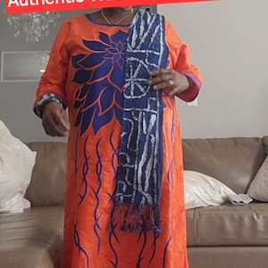 K&ouml;nnte beinhalten: Ein orangefarbenes Kleid mit blauen Blumen- und rankenartigen Mustern. Das Kleid wird mit einem passenden Kopftuch und einem blau-wei&szlig;en Schal getragen. Der Text "Authentic Washable Ndop Scarf" ist im Bild zu sehen.
