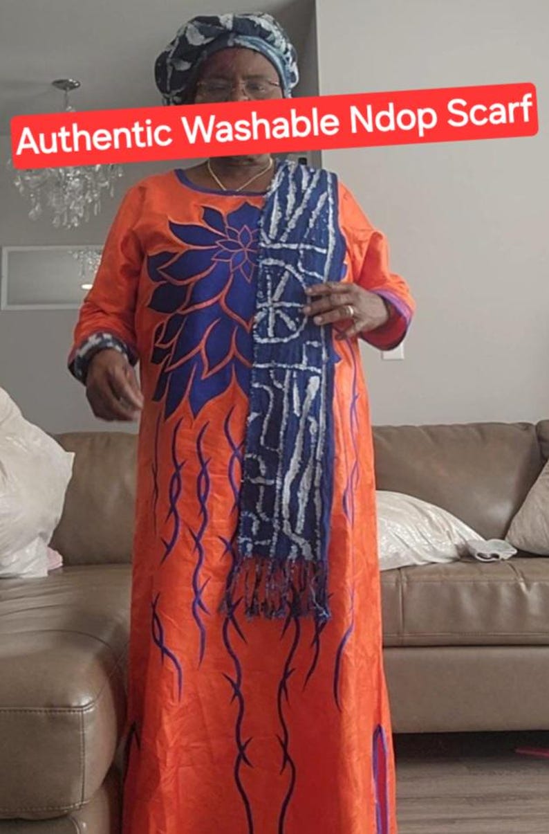 K&ouml;nnte beinhalten: Ein orangefarbenes Kleid mit blauen Blumen- und rankenartigen Mustern. Das Kleid wird mit einem passenden Kopftuch und einem blau-wei&szlig;en Schal getragen. Der Text "Authentic Washable Ndop Scarf" ist im Bild zu sehen.