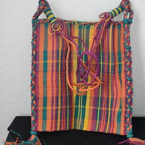May include: A colorful woven bag with vertical stripes in orange, yellow, purple, and teal. The bag has braided edges and handles, with tassels at the bottom. The handles are teal and yellow, and the tassels are teal, orange, and purple.