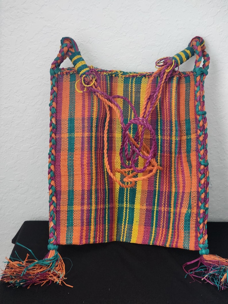 May include: A colorful woven bag with vertical stripes in orange, yellow, purple, and teal. The bag has braided edges and handles, with tassels at the bottom. The handles are teal and yellow, and the tassels are teal, orange, and purple.