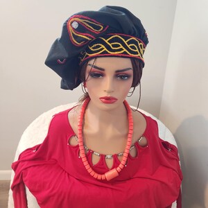 May include: A black fabric hat with intricate yellow and red embroidery, paired with a red top and a coral-colored beaded necklace. The hat features a decorative design around the brim and on the crown.