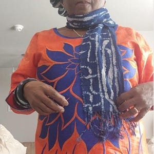 K&ouml;nnte beinhalten: Ein orangefarbenes Kleid mit blauen Blumenstickereien und langen &Auml;rmeln. Das Kleid wird mit einem blau-wei&szlig; gemusterten Schal und einem passenden Hut kombiniert. Das Outfit wird von einer Person getragen.