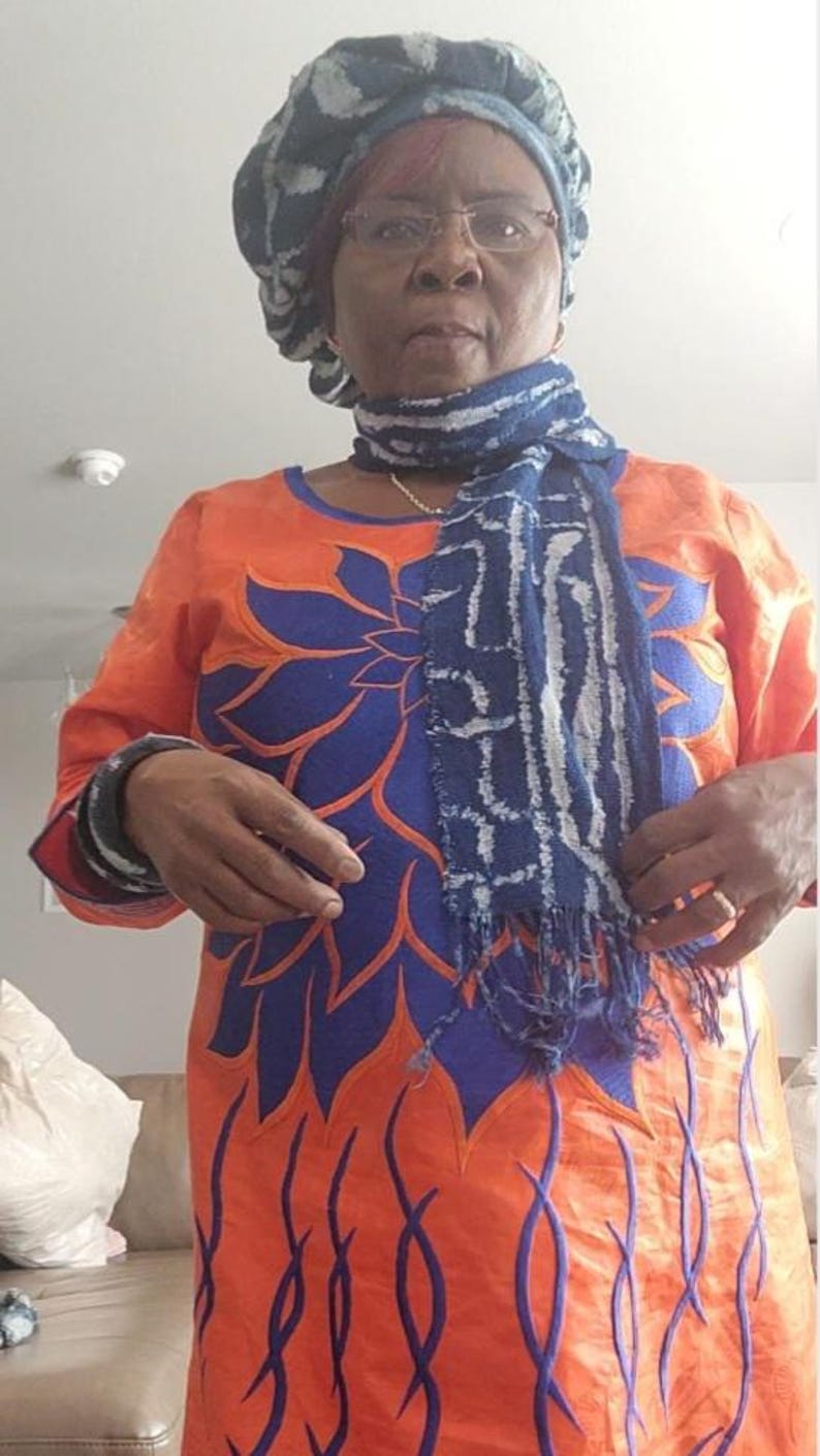 K&ouml;nnte beinhalten: Ein orangefarbenes Kleid mit blauen Blumenstickereien und langen &Auml;rmeln. Das Kleid wird mit einem blau-wei&szlig; gemusterten Schal und einem passenden Hut kombiniert. Das Outfit wird von einer Person getragen.