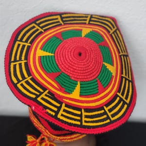 May include: A handmade, colorful hat with a circular design. The hat features a red center with green and yellow accents, surrounded by black and yellow geometric patterns. A tassel hangs from the side. The hat is likely made of woven or knitted materials.