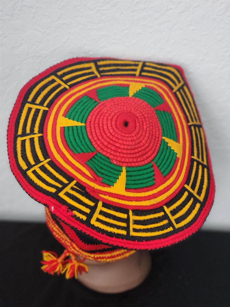 May include: A handmade, colorful hat with a circular design. The hat features a red center with green and yellow accents, surrounded by black and yellow geometric patterns. A tassel hangs from the side. The hat is likely made of woven or knitted materials.