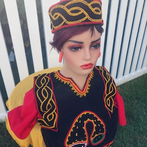 Pu&ograve; includere: Un manichino indossa un completo ricamato nero e rosso, compresi cappello e orecchini abbinati. L'outfit presenta cuciture decorative dorate e rosse. Il manichino &egrave; posizionato davanti a una recinzione bianca e erba verde.