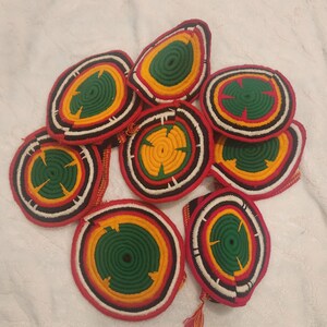 May include: A collection of handmade, circular coasters. Each coaster features a vibrant design with concentric rings of red, black, white, yellow, and green. The coasters are made of a woven material, and some have decorative tassels.