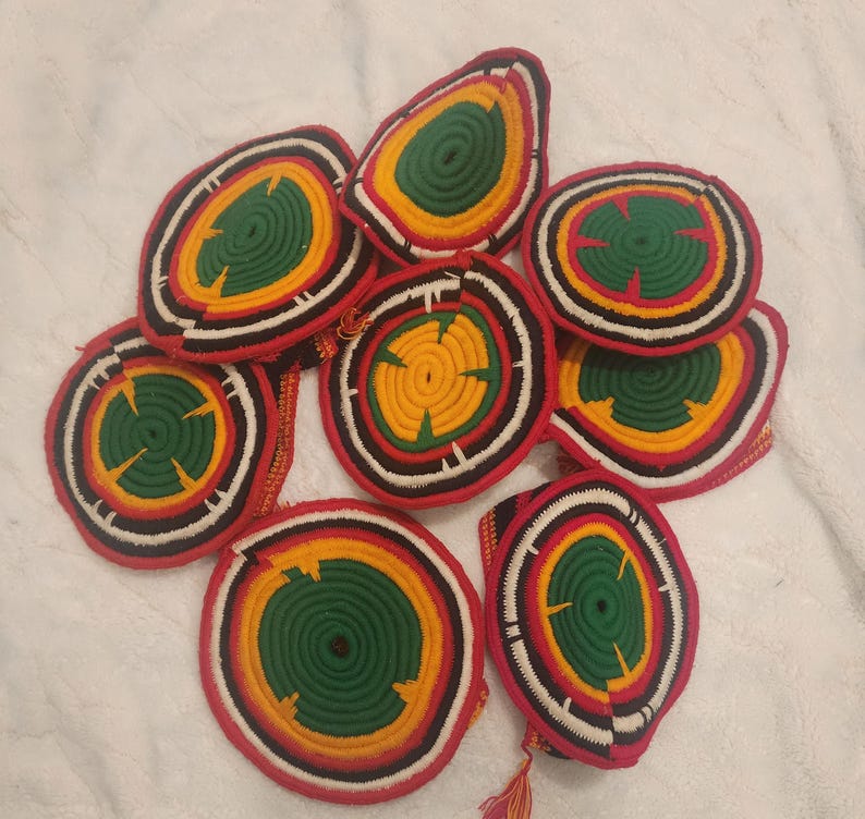 May include: A collection of handmade, circular coasters. Each coaster features a vibrant design with concentric rings of red, black, white, yellow, and green. The coasters are made of a woven material, and some have decorative tassels.
