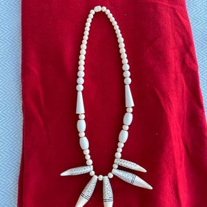 K&ouml;nnte beinhalten: Eine wei&szlig;e Perlenkette mit einem Anh&auml;nger aus f&uuml;nf wei&szlig;en, spitzen, geschnitzten Perlen mit schwarzen Markierungen. Die Kette liegt auf einem roten Stoffhintergrund.