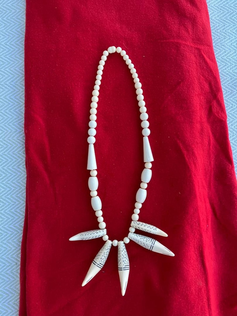 K&ouml;nnte beinhalten: Eine wei&szlig;e Perlenkette mit einem Anh&auml;nger aus f&uuml;nf wei&szlig;en, spitzen, geschnitzten Perlen mit schwarzen Markierungen. Die Kette liegt auf einem roten Stoffhintergrund.
