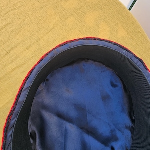 Pu&ograve; includere: Primo piano dell'interno di un cappello. Presenta una fodera in raso blu navy, una fascia elastica nera e un bordo rosso e nero. Il cappello &egrave; appoggiato su una superficie gialla.