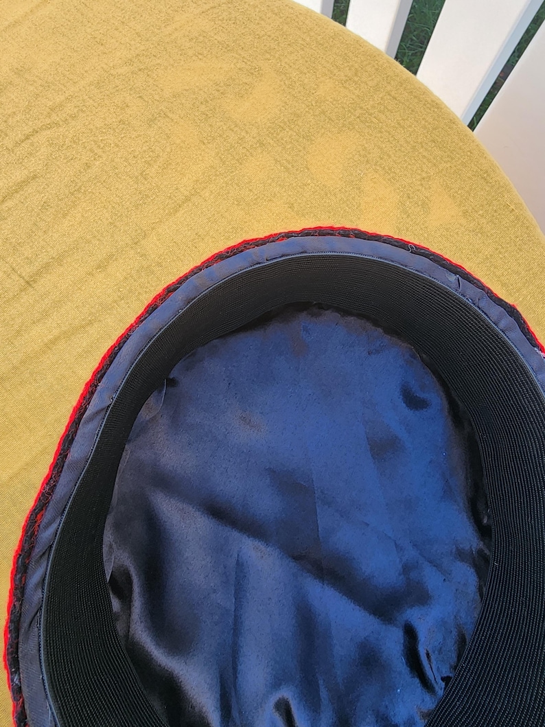 Pu&ograve; includere: Primo piano dell'interno di un cappello. Presenta una fodera in raso blu navy, una fascia elastica nera e un bordo rosso e nero. Il cappello &egrave; appoggiato su una superficie gialla.
