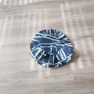 Puede incluir: Un sombrero redondo con un estampado de rayas azules y blancas y un borde con volantes. El sombrero presenta l&iacute;neas blancas sobre una tela azul oscuro. El sombrero es de tela y est&aacute; sobre una superficie de madera.