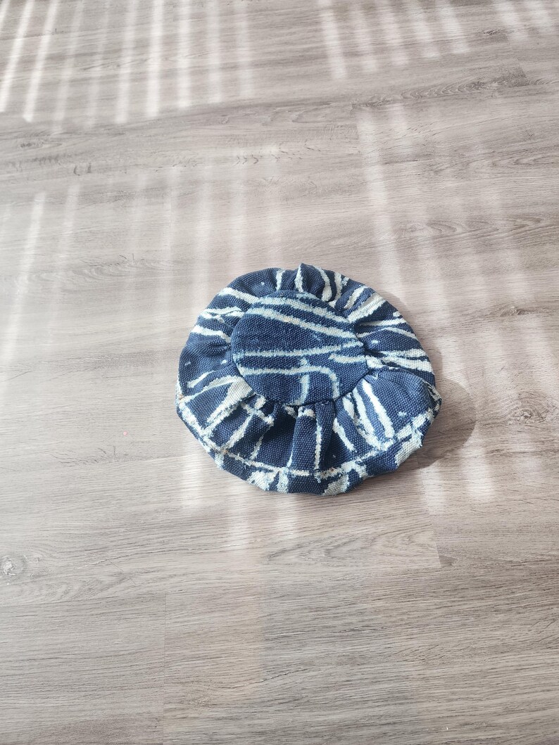 Puede incluir: Un sombrero redondo con un estampado de rayas azules y blancas y un borde con volantes. El sombrero presenta l&iacute;neas blancas sobre una tela azul oscuro. El sombrero es de tela y est&aacute; sobre una superficie de madera.