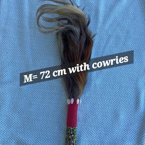 May include: A decorative wand with a handle wrapped in red fabric and adorned with colorful beads. The top features a bundle of brown, gray, and tan hair. The text "M= 72 cm with cowries" is visible.