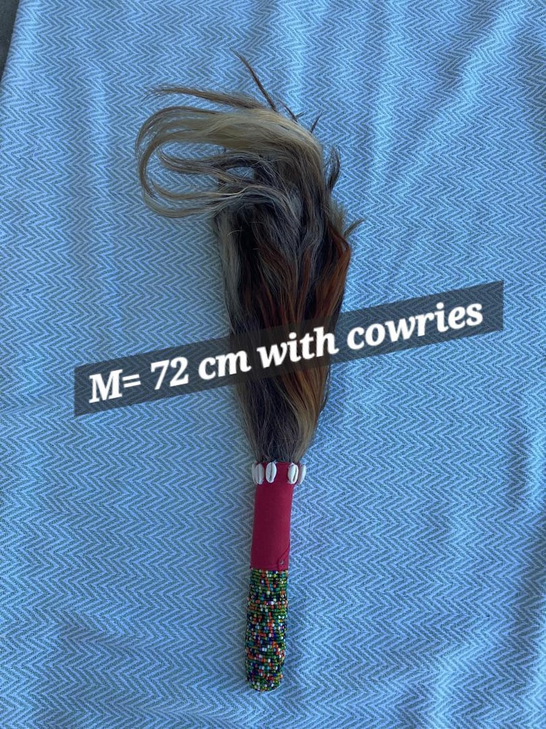 May include: A decorative wand with a handle wrapped in red fabric and adorned with colorful beads. The top features a bundle of brown, gray, and tan hair. The text "M= 72 cm with cowries" is visible.