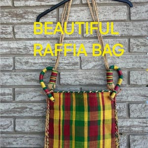 May include: A raffia bag with a plaid pattern in red, yellow, and green. The bag has rope handles and the text "BEAUTIFUL RAFFIA BAG" in yellow. The bag is hanging on a brick wall.