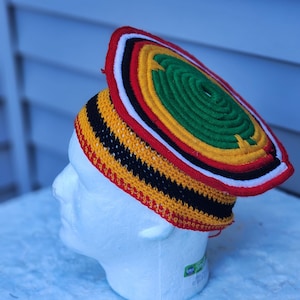 May include: A colorful, handmade hat with a circular top and a brim. The hat features a vibrant design with red, yellow, black, white, and green colors. The hat is displayed on a white mannequin head.