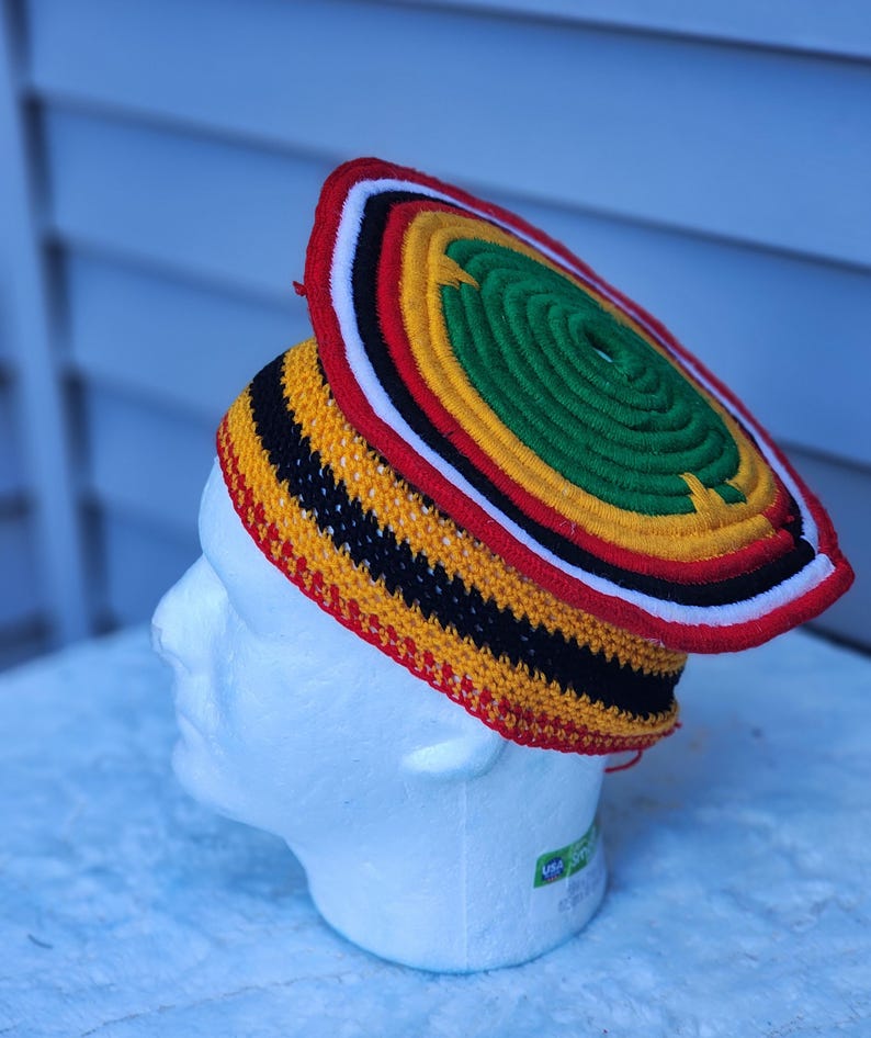 May include: A colorful, handmade hat with a circular top and a brim. The hat features a vibrant design with red, yellow, black, white, and green colors. The hat is displayed on a white mannequin head.