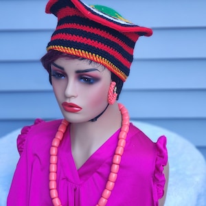 May include: A mannequin wearing a vibrant, striped hat in red, black, and yellow, paired with matching coral-colored jewelry and a bright pink top. The hat features a unique, curved brim and a colorful design on top. The jewelry includes hoop earrings and a long beaded necklace.