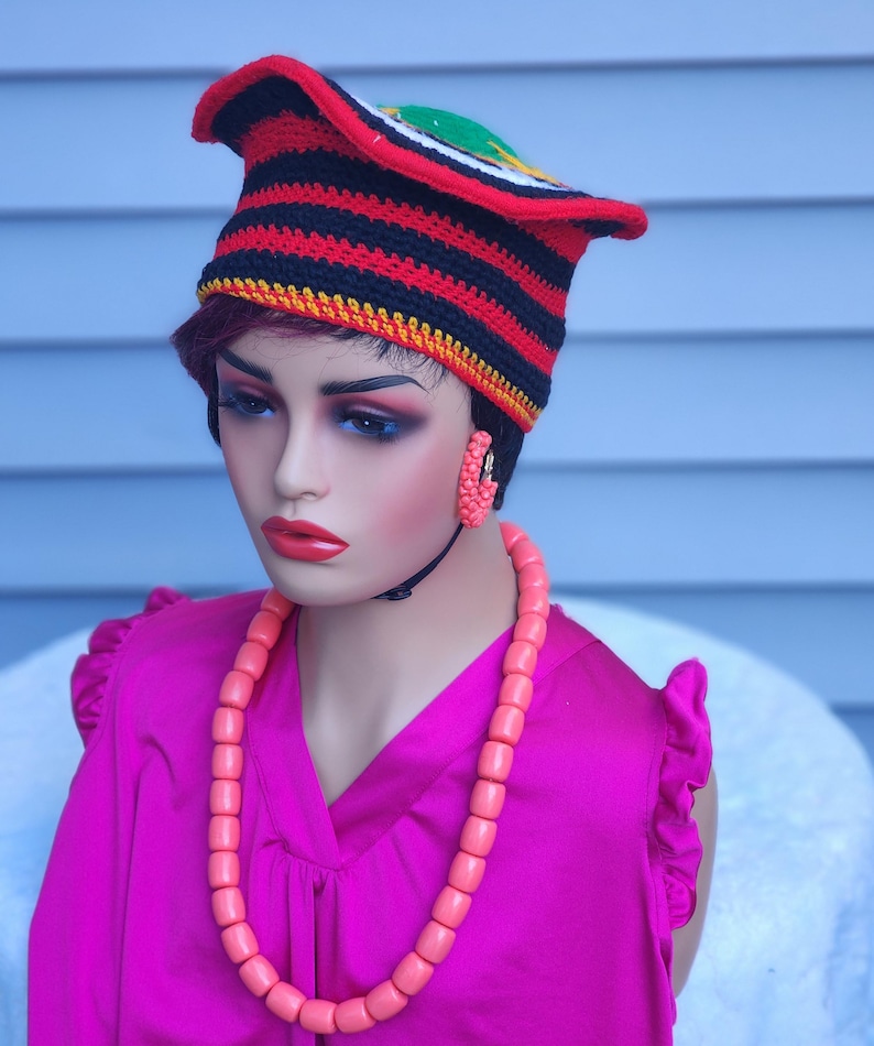 May include: A mannequin wearing a vibrant, striped hat in red, black, and yellow, paired with matching coral-colored jewelry and a bright pink top. The hat features a unique, curved brim and a colorful design on top. The jewelry includes hoop earrings and a long beaded necklace.
