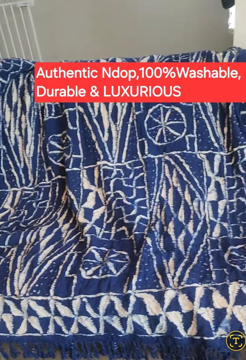 K&ouml;nnte beinhalten: Blau-wei&szlig; gemusterter Stoff mit Fransen am unteren Rand. Der Stoff weist ein sich wiederholendes Design aus geometrischen Formen und Linien auf. Der Text "Authentic Ndop, 100% Washable, Durable & LUXURIOUS" wird in einem roten Banner angezeigt.
