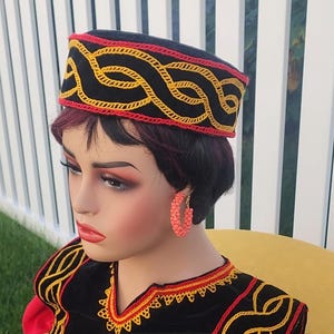 Pu&ograve; includere: Un cappello nero con dettagli ricamati rossi e gialli. Il cappello presenta un design intrecciato ed &egrave; indossato da un manichino. Il manichino indossa anche un top ricamato nero e rosso e orecchini a cerchio color corallo. Lo sfondo &egrave; una recinzione bianca.