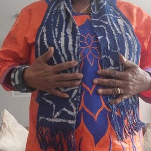 K&ouml;nnte beinhalten: Ein orangefarbenes Kleid mit blauen und orangefarbenen Blumenstickereien. Ein blauer und wei&szlig;er Schal mit Fransen ist &uuml;ber dem Kleid drapiert. Die Person tr&auml;gt eine passende Kopfbedeckung und Armb&auml;nder.