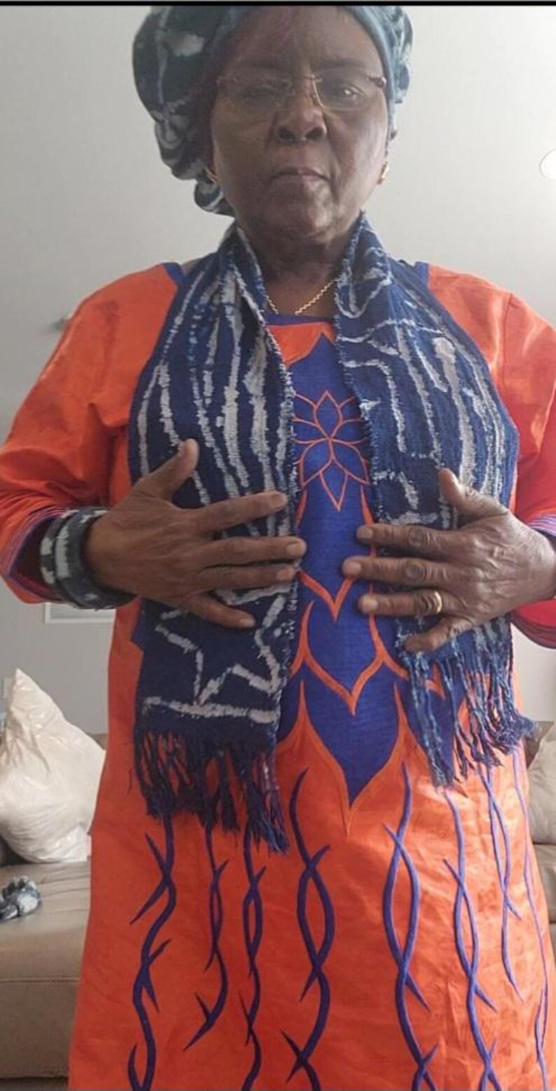 K&ouml;nnte beinhalten: Ein orangefarbenes Kleid mit blauen und orangefarbenen Blumenstickereien. Ein blauer und wei&szlig;er Schal mit Fransen ist &uuml;ber dem Kleid drapiert. Die Person tr&auml;gt eine passende Kopfbedeckung und Armb&auml;nder.