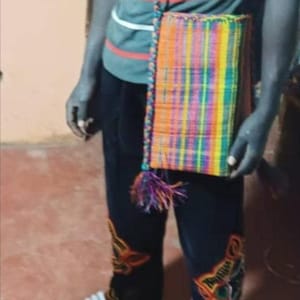 May include: A rectangular, woven shoulder bag with a vibrant, multicolored pattern of orange, yellow, green, blue, and purple. The bag has a matching strap and a tassel. The person is wearing a striped shirt and dark pants.