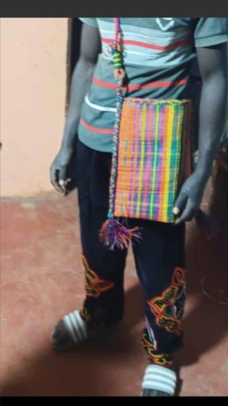 May include: A rectangular, woven shoulder bag with a vibrant, multicolored pattern of orange, yellow, green, blue, and purple. The bag has a matching strap and a tassel. The person is wearing a striped shirt and dark pants.