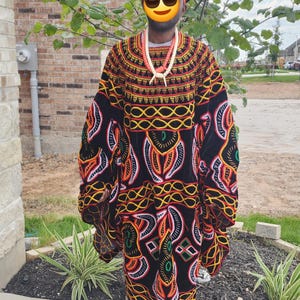May include: A colorful, patterned African robe with long sleeves. The robe features a black background with orange, yellow, and white geometric designs. A matching head wrap and beaded necklace complete the ensemble. The person is wearing blue shoes.