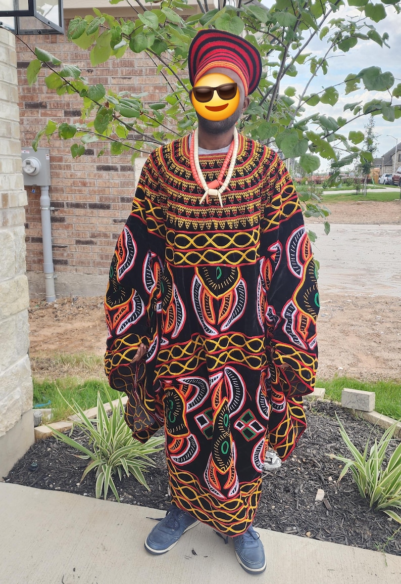 May include: A colorful, patterned African robe with long sleeves. The robe features a black background with orange, yellow, and white geometric designs. A matching head wrap and beaded necklace complete the ensemble. The person is wearing blue shoes.