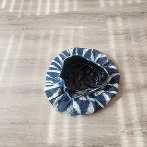 Puede incluir: Un gorro de boina con estampado azul y blanco. El sombrero presenta un exterior azul oscuro con patrones triangulares blancos que irradian desde el centro. El interior es negro. El sombrero est&aacute; sobre una superficie de madera clara.