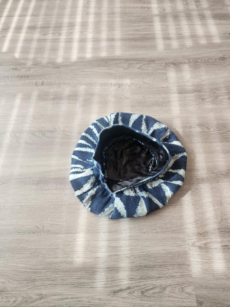 Puede incluir: Un gorro de boina con estampado azul y blanco. El sombrero presenta un exterior azul oscuro con patrones triangulares blancos que irradian desde el centro. El interior es negro. El sombrero est&aacute; sobre una superficie de madera clara.