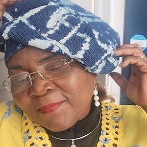 Puede incluir: Un sombrero con estampado azul y blanco. El sombrero se usa con una chaqueta amarilla con ribetes azules y amarillos, un collar plateado con un colgante de piedra blanca y aretes de piedra blanca. La persona lleva gafas.