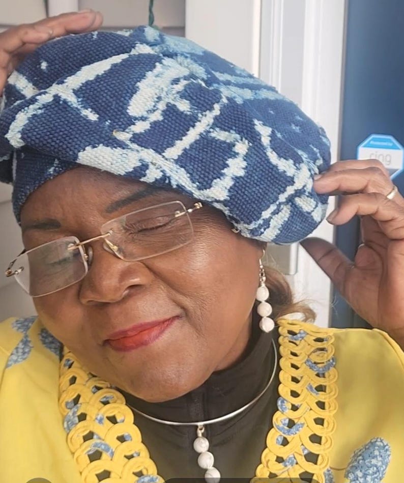 Puede incluir: Un sombrero con estampado azul y blanco. El sombrero se usa con una chaqueta amarilla con ribetes azules y amarillos, un collar plateado con un colgante de piedra blanca y aretes de piedra blanca. La persona lleva gafas.