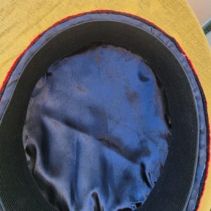 Pu&ograve; includere: Vista interna di un cappello con un elastico nero e una fodera in raso blu. Il cappello ha un bordo rosso e un bordo esterno nero. Il cappello &egrave; posto su uno sfondo giallo.