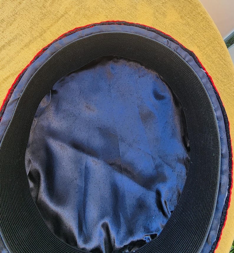 Pu&ograve; includere: Vista interna di un cappello con un elastico nero e una fodera in raso blu. Il cappello ha un bordo rosso e un bordo esterno nero. Il cappello &egrave; posto su uno sfondo giallo.