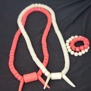 May include: Two necklaces and a bracelet. One necklace is coral, the other is white. The bracelet alternates coral and white beads. The necklaces have a cylindrical center piece and pointed ends. The background is black.