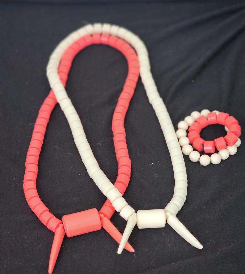 May include: Two necklaces and a bracelet. One necklace is coral, the other is white. The bracelet alternates coral and white beads. The necklaces have a cylindrical center piece and pointed ends. The background is black.