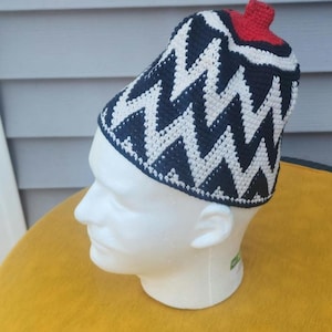 May include: A handmade crochet hat with a zig-zag pattern in navy blue and white. The hat has a red accent on top and is displayed on a white mannequin head. The hat is likely a winter accessory.