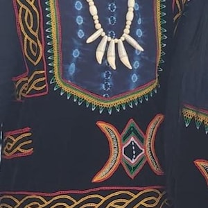 K&ouml;nnte beinhalten: Ein schwarz-blaues gemustertes Hemd mit aufwendigen goldenen Stickereien. Das Hemd hat ein gro&szlig;es, farbenfrohes geometrisches Design auf der Vorderseite und eine wei&szlig;e Perlenkette mit Knochenanh&auml;ngern.