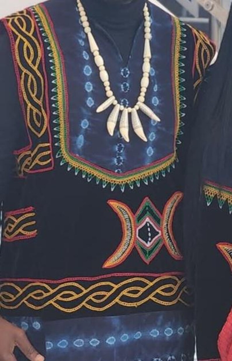 K&ouml;nnte beinhalten: Ein schwarz-blaues gemustertes Hemd mit aufwendigen goldenen Stickereien. Das Hemd hat ein gro&szlig;es, farbenfrohes geometrisches Design auf der Vorderseite und eine wei&szlig;e Perlenkette mit Knochenanh&auml;ngern.