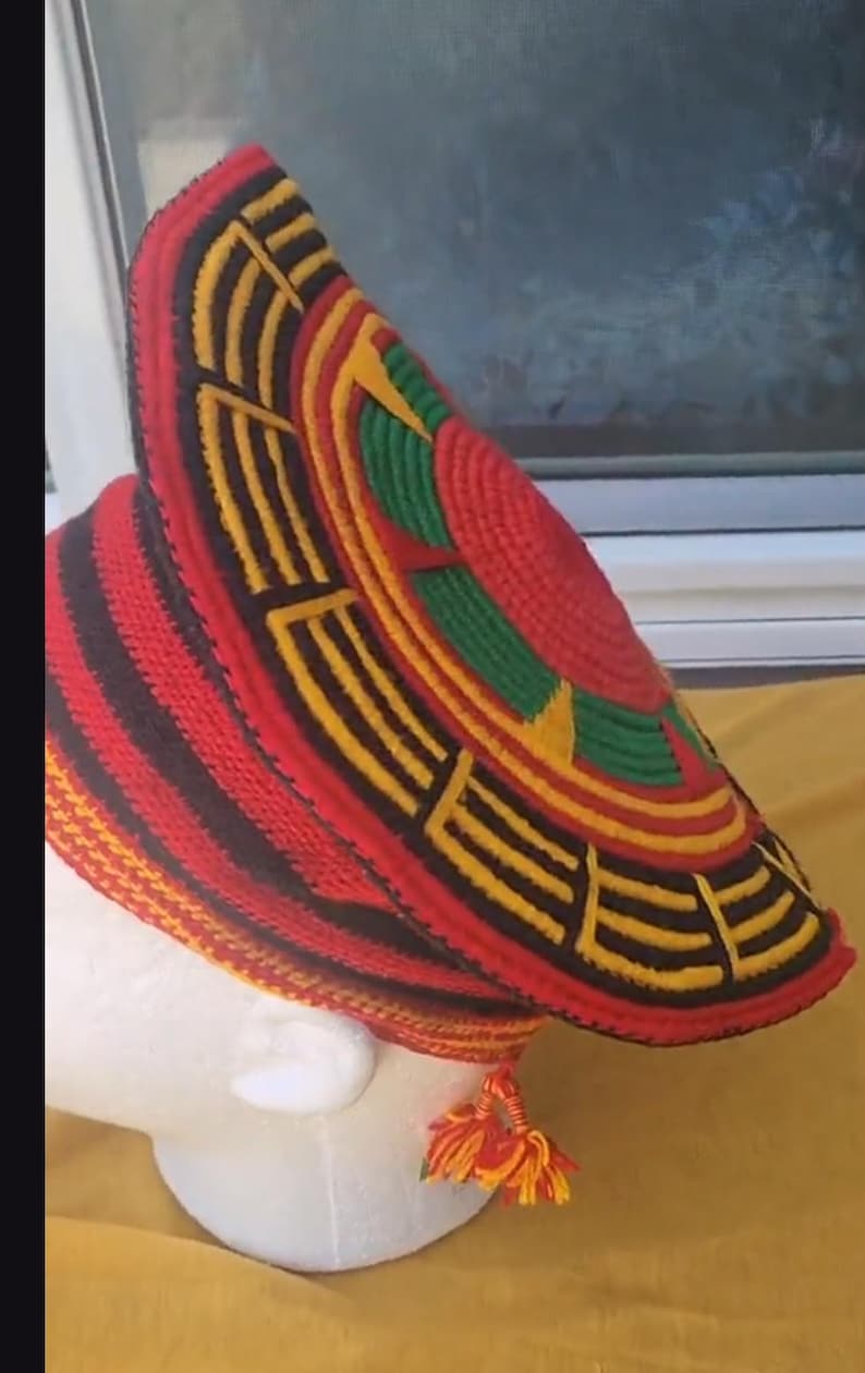May include: A vibrant, handmade hat with a wide brim, featuring a striking pattern of red, black, yellow, and green. The hat has a unique, textured design and a tassel detail. This headwear is a bold statement piece.