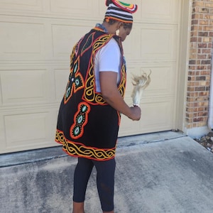 May include: A person wearing a black vest with colorful embroidery, a matching hat, and red shoes. The vest features intricate patterns in red, yellow, and green on a black background. The hat has a striped design with a curved top. The person is holding a white object.